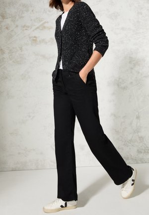 Czarny dziergany kardigan z białymi kropkami, biała koszulka i czarne spodnie, połączone z białymi trampkami z czarnymi akcentami, pozycja stojąca.