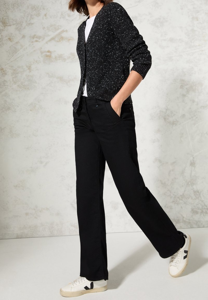 Czarny dziergany kardigan z białymi kropkami, biała koszulka i czarne spodnie, połączone z białymi trampkami z czarnymi akcentami, pozycja stojąca.