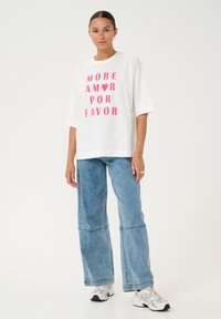 Camiseta blanca de gran tamaño con un atrevido texto en rosa que dice "MORE AMOR POR FAVOR." Combinada con unos jeans de denim azul de pierna ancha y zapatillas plateadas.