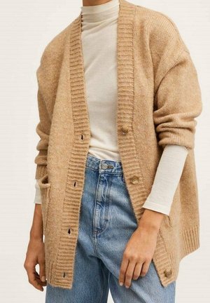 Osoba w beżowym kardiganie na guziki, nosząca pod spodem biały top z długim rękawem oraz niebieskie, wysokowjaśniane dżinsy, z rękami swobodnie opuszczonymi wzdłuż ciała.