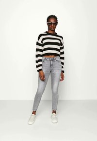 Pull court rayé noir et blanc, jean gris ajusté et baskets blanches. Le modèle porte des lunettes de soleil. Tissu lisse, coupe ajustée, style décontracté.