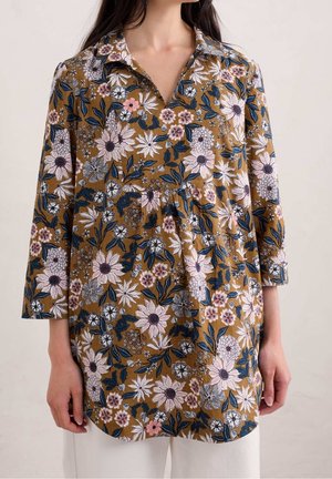 Kvinde iført en brun tunika med blomsterprint, trekvartærmer og krave, kombineret med hvide bukser.