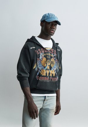 Homme portant un sweat à capuche de sport noir délavé avec texte graphique et une casquette en jean bleu clair, debout devant un fond gris uni.