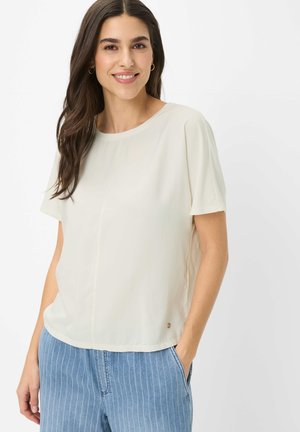 Femme souriante aux longs cheveux foncés portant un chemisier blanc ample à manches courtes et un pantalon bleu à fines rayures, debout avec une main dans la poche.