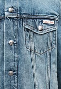 Φως μπλε τζιν μπουφάν μπροστά με μεταλλικά κουμπιά και τσέπη με καπάκι που φέρει κόκκινη και λευκή ετικέτα Calvin Klein.