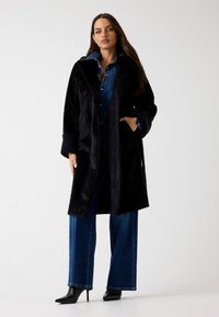 Schwarzer Kunstpelzmantel mit Kapuze, offenem Vorderteil und gerippten Ärmeln, kombiniert mit einem Jeanshemd und schwarzen High-Heels.