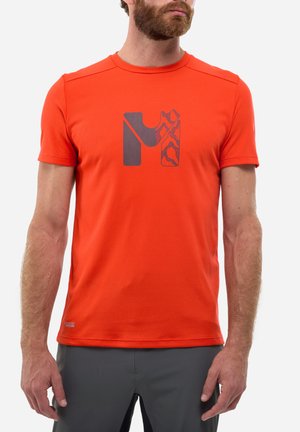 Mann trägt ein leuchtend orangefarbenes, kurzärmeliges Sportshirt mit dunklem grauem "M"-Grafik auf der Brust, kombiniert mit dunkelgrauen Hosen.