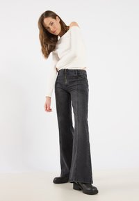 Vaqueros negros de campana con detalles de costura y cierre de cremallera, combinados con una blusa blanca ajustada de manga larga. Textura de tela suave.