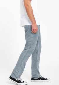 Jeans de denim azul claro con un ajuste recto, que presentan bolsillos cosidos y un pequeño detalle de logo. Llevados con zapatillas negras.