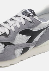 Sneaker grigio realizzato in materiali scamosciati e mesh, con un accento del logo nero e dettagli perforati sulla punta, lacci bianchi e una suola leggera.