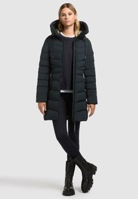 Cappotto imbottito verde scuro, di lunghezza media, con cappuccio e chiusura a zip. Presenta una texture trapuntata e tasche laterali, abbinato a leggings neri e stivali.