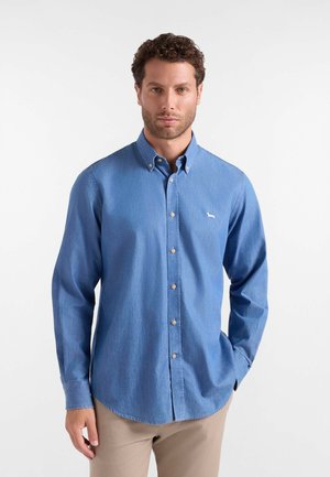 Uomo che indossa una camicia blu con bottoni con un piccolo logo bianco sul petto e pantaloni beige, in piedi contro uno sfondo bianco uniforme.