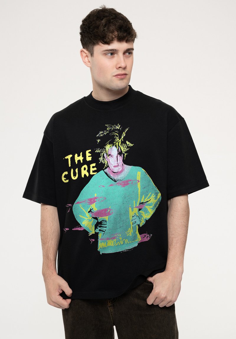 Black oversized t-shirt featuring a colorful graphic design of a figure, with yellow text "THE CURE"; short sleeves and a round neckline.