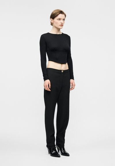 Femme aux cheveux courts portant un haut court noir à manches longues, un pantalon noir taille haute et des bottines à talons noires sur un fond blanc.