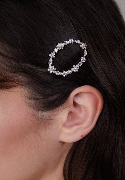 Barrette en argent de forme ovale, ornée de petites fleurs artificielles et d'accents scintillants, fixée dans des cheveux noirs près de l'oreille.
