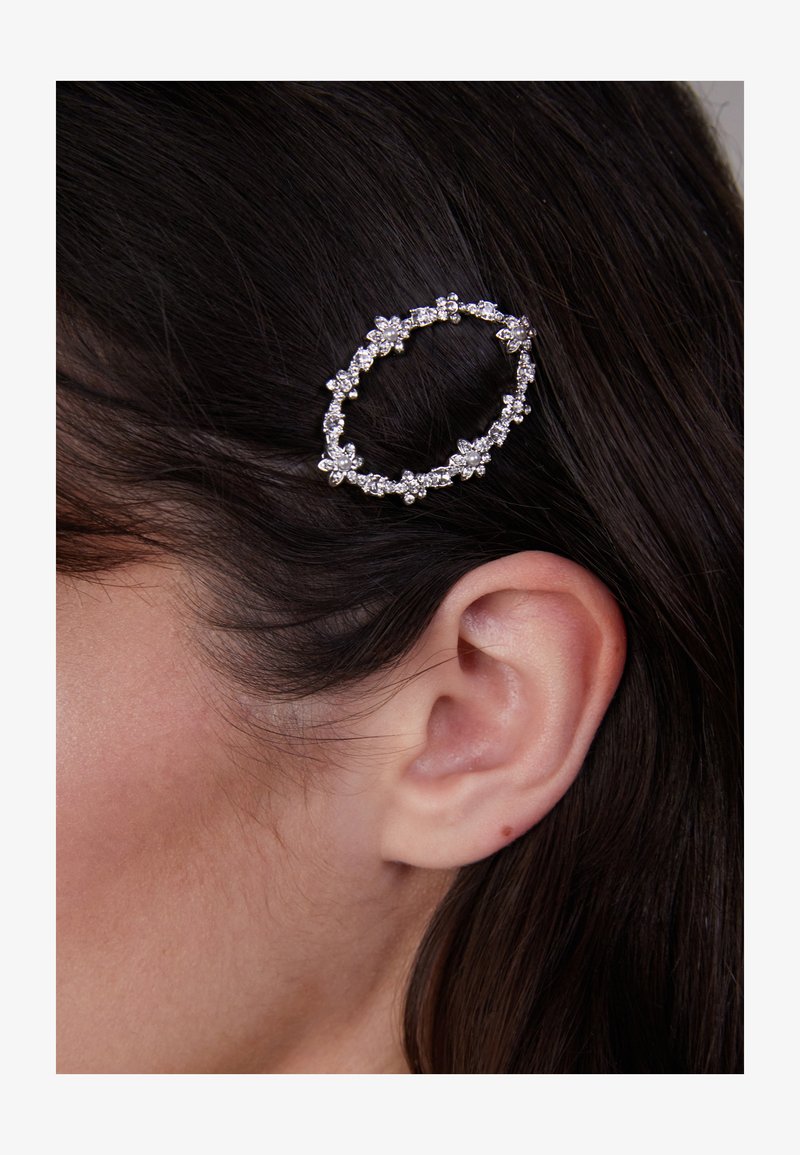 Auksinės spalvos plaukų segtukas ovalo formos, puošnus mažomis dirbtinėmis gėlėmis ir žėrinčiomis detalėmis, prispaustas prie tamsių plaukų šalia ausies.