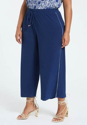 Femme portant un pantalon large bleu marine avec une bande blanche sur le côté et des sandales à talons hauts crème avec des liens à la cheville et des breloques.