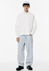 Camicia bianca oversize con spalle scese, jeans ampi blu chiaro e scarpe chunky nere. Un berretto grigio completa l'outfit.