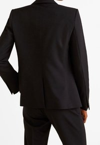 Personne portant une veste de costume noire ajustée et un pantalon assorti, vue de dos sur un fond blanc uni.