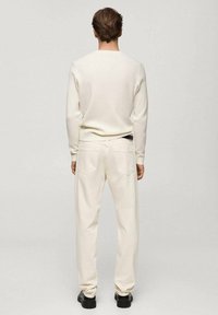 White ribbed long-sleeve sweater paired with light-colored trousers, featuring back pockets and textured fabric, worn with black shoes.