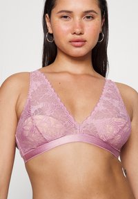 Sutiã de renda em rosa suave com forma triangular, bordas em forma de ondas e faixa larga em cetim. Padrão floral transparente cobre o tecido.