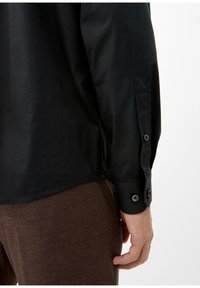 Camisa negra de manga larga con puños abotonados y textura suave. Pantalones marrones texturizados visibles por la parte inferior. Diseño simple sin patrones.