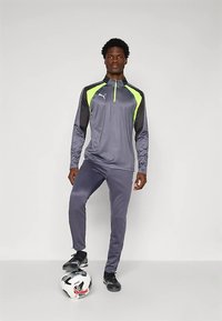 Egy férfi szürke és neon zöld Puma sportöltözetben áll, egyik lába egy fehér és fekete focilabdán, egy sima háttér előtt.