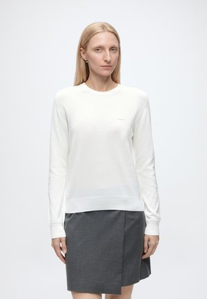 Donna bionda che indossa un maglione bianco a maniche lunghe e una gonna grigio scuro al ginocchio, in piedi davanti a uno sfondo bianco semplice.