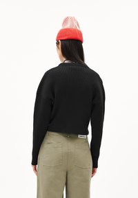 Pull en maille côtelée noir avec un design raccourci et des manches longues, associé à un pantalon vert olive avec des poches arrière et une étiquette visible.