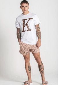 Bílé tričko s velkým hnědým logem "K", vzorované béžové kraťasy a hnědé pantofle. Zahrnuje viditelné tetování na pažích a nohách.