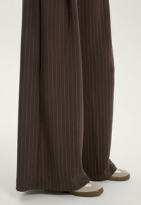 Wijdbenige broeken in donkerbruin met subtiele verticale streepjes, gecombineerd met tan en grijze schoenen met een ronde neus en contrasterende accenten.