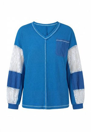 Chemise bleue à manches longues avec col en V, poche avant et panneaux en dentelle blanche sur les manches alternant avec un tissu bleu uni.