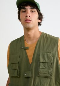 Πράσινο αμάνικο γιλέκο με πολλαπλές μπροστινές τσέπες, η υφή του υφάσματος είναι λεία, φορεμένο πάνω από ένα κίτρινο πουκάμισο, με κουμπώματα snap και απλό σχέδιο.