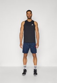 Zwarte sportieve tanktop met wit logo, gecombineerd met marineblauwe shorts met rode logoprints. Zwarte sneakers en gestreepte sokken maken de look compleet.