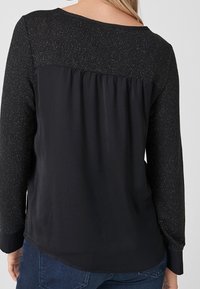 Schwarze Bluse mit einer strukturierten, glitzernden oberen Partie, langen Ärmeln und einem leicht geschwungenen Saum. Weicher Stoff mit einer lockeren Passform.