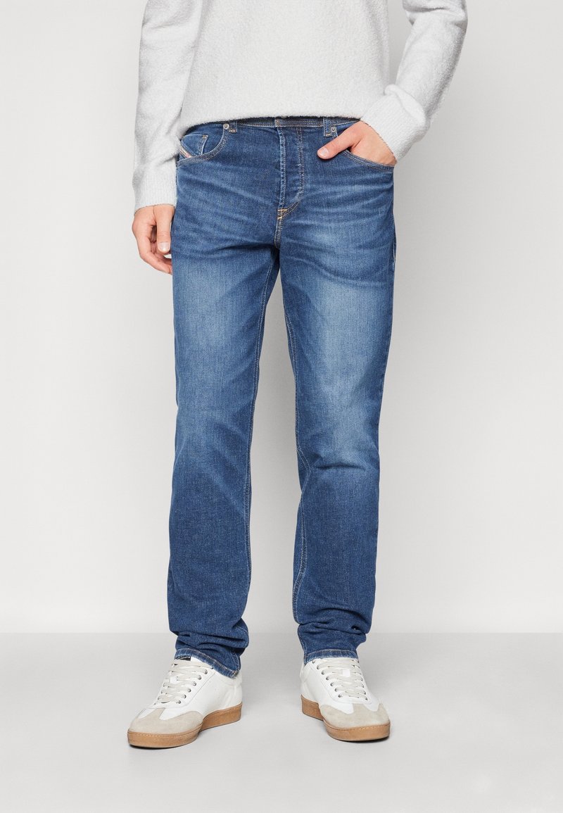 Jeans in denim con un lavaggio blu medio, taglio dritto, design a cinque tasche e dettagli di cucitura sottili, abbinati a sneaker bianche.