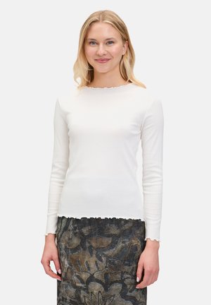 Haut à manches longues blanc avec un col et un ourlet festonnés, fabriqué en tissu doux, associé à une jupe foncée à motifs avec des designs floraux.