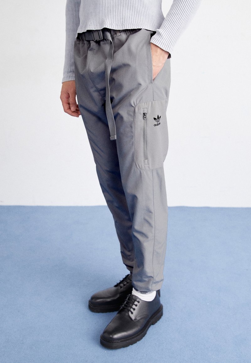 Šedé cargo kalhoty s elastickým pasem, šňůrkou na stažení a boční kapsou na zip. Černý logo akcent na stehně. Stylizováno s černými botami.