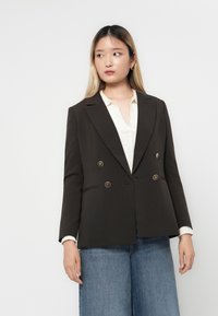 Brun dobbeltspent blazer med gullknappeaksenter, nothals, og strukturerte skuldre, brukt over en hvit strikket topp og vidt legget jeans.