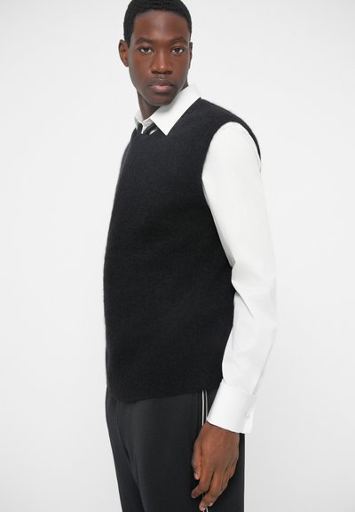 Čierny huňatý vest z mäkkého materiálu, s uvoľneným strihom a bezrukávovým dizajnom, nosený cez bielu košeľu s golierom.