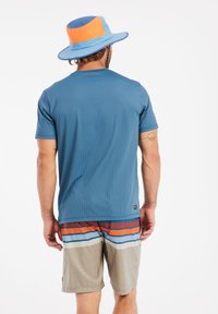 Blaues Kurzarmhemd mit gerippter Struktur, kombiniert mit gestreiften Shorts in Orange, Blau, Weiß und Braun. Eine blaue und orangefarbene Mütze rundet den Look ab.
