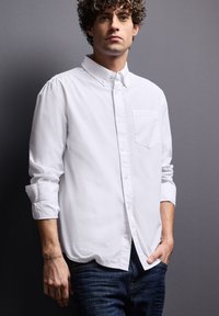 Witte overhemdbloes met een enkele borstzak, lange mouwen opgerold bij de manchetten, een gladde textuur, gedragen over donkere spijkerbroeken.