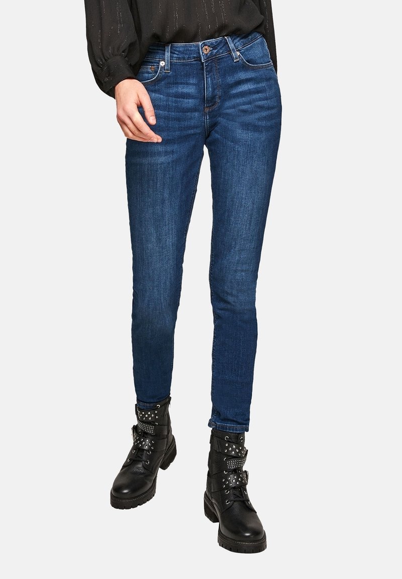Jean skinny en denim bleu foncé à texture lisse, avec une conception classique à cinq poches et une taille mi-haute, associé à des bottines noires cloutées.