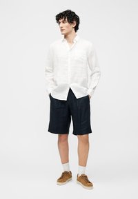 Άνδρας με σγουρά μαλλιά που φοράει λευκό πουκάμισο με μακριά μανίκια, σκούρα σορτς μέχρι το γόνατο, λευκές κάλτσες και καφέ αθλητικά παπούτσια, στέκεται με τα χέρια στις τσέπες.