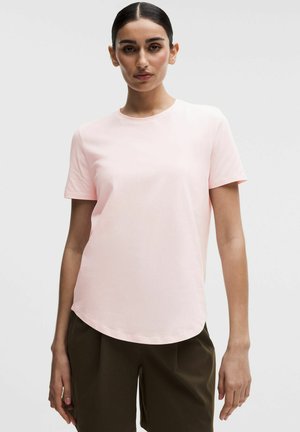 Kobieta o ciemnych włosach, ubrana w jasnoróżowy t-shirt z krótkim rękawem i ciemnobrązowe spodnie, stojąca na tle jednolitego jasnego tła.