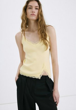 Mujer con cabello largo y ondulado, vestida con un camisón amarillo con encaje y pantalones plisados negros, de pie con una mano en el bolsillo.