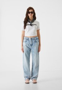 Camiseta corta blanca con texto negro, jeans de pierna ancha azul claro, zapatillas rosa claro y una bufanda negra con patrón alrededor del cuello.