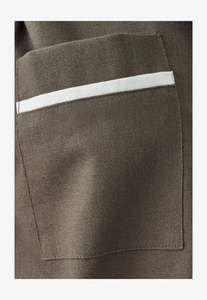 Brauner Stoff mit einer strukturierten Oberfläche hat eine rechteckige Tasche, die an der oberen Kante mit einem weißen horizontalen Streifen versehen ist.