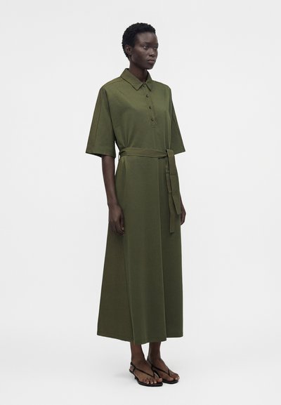 Γυναίκα στέκεται φορώντας ένα μεσάτο φόρεμα ελιάς με κουμπιά, γιακά, κοντά μανίκια και ζώνη στη μέση δεμένη, σε συνδυασμό με μαύρα σανδάλια.
