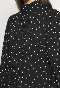 Blusa nera con pois bianchi, dotata di un colletto alto e un riporto annodato sulla parte anteriore. Il tessuto presenta una superficie texturizzata e una vestibilità comoda.